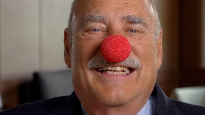 An older man with a mustache smiles warmly while wearing a red clown nose, dressed in a suit, with a blurred indoor background.