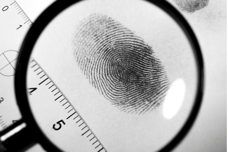 A magnifying glass enlarges a fingerprint on white paper, with a ruler next to the print for scale. Another faint fingerprint is visible in the background.