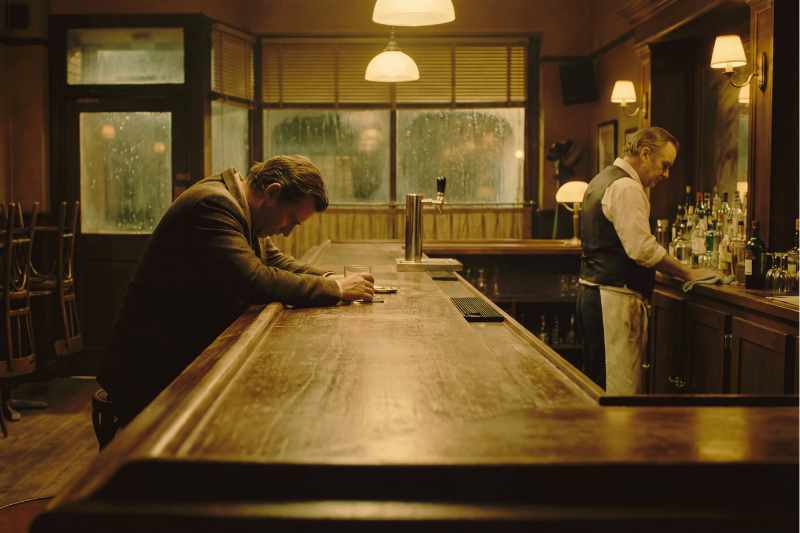 A man leans over a wooden bar with his head down, holding a glass, while a bartender stands behind the counter arranging bottles. The dimly lit bar appears empty, with rain visible outside the windows.