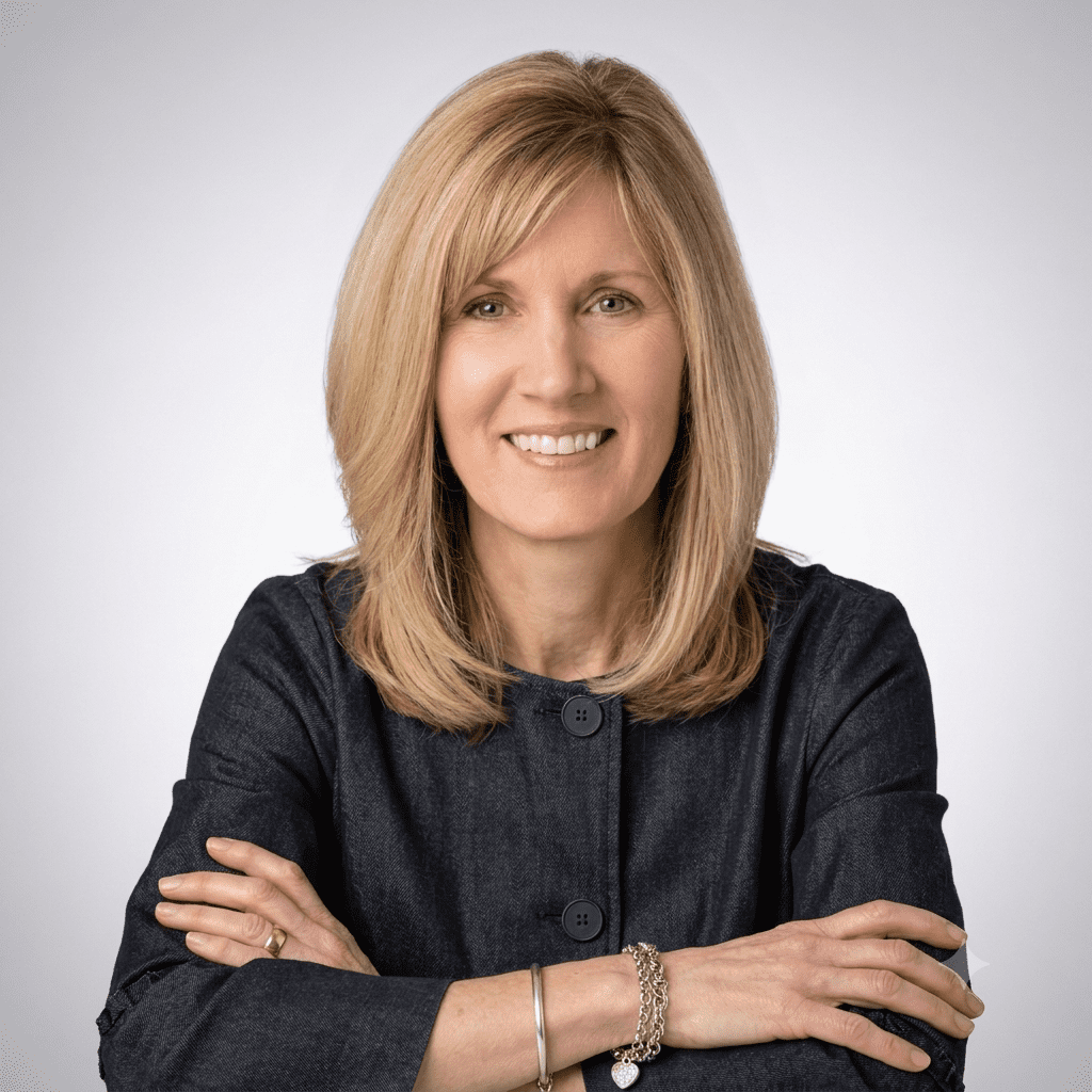 A woman with straight blonde hair, wearing a dark jacket, sits with her arms crossed and smiles at the camera against a plain light background.