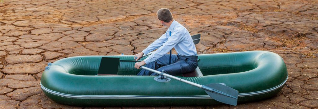 A man in business attire sits in an inflatable boat on dry, cracked ground, holding a laptop and an oar, looking over his shoulder.