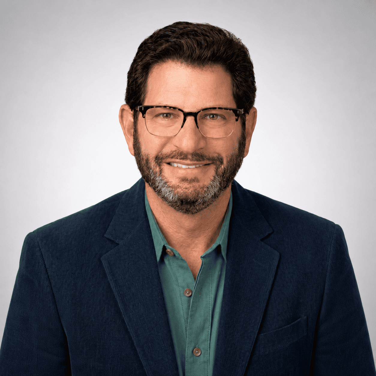 A man with short dark hair, beard, and glasses, wearing a green shirt and blue blazer, smiles in a professional studio portrait with a light gray background.