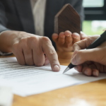 A person points to a section of a contract while another person signs it with a pen; a small wooden house model is held in the background, suggesting a real estate agreement.