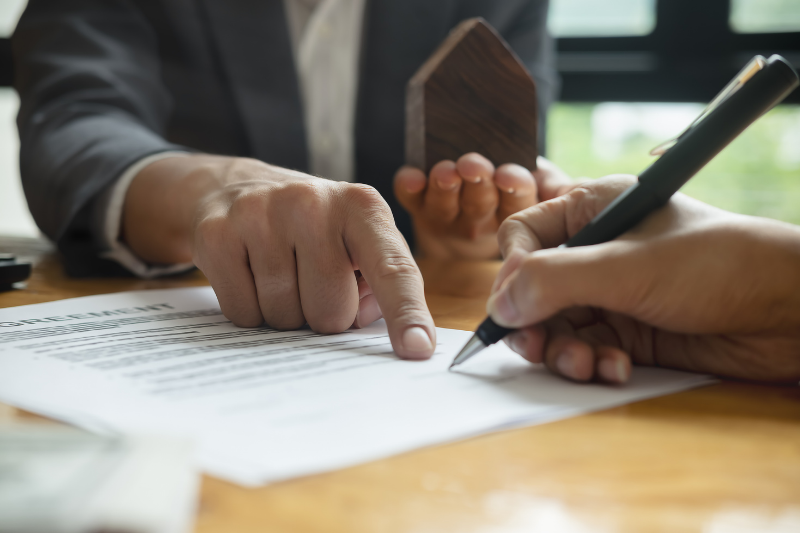 A person points to a section of a contract while another person signs it with a pen; a small wooden house model is held in the background, suggesting a real estate agreement.
