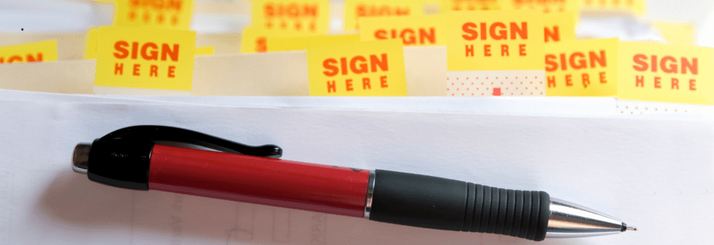 A red and black pen rests on a stack of papers, which are marked with multiple yellow Sign Here sticky tabs sticking out from the edges.