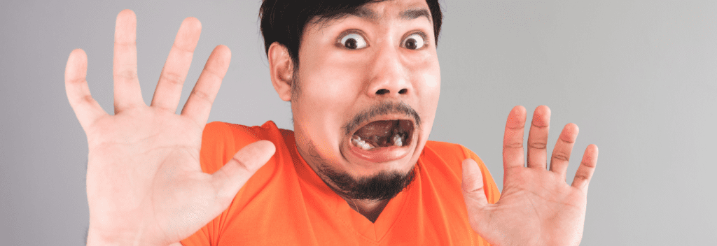 A man in an orange shirt looks frightened, eyes wide and mouth open in shock, with both hands raised in front of him as if to protect himself. The background is plain and light-colored.