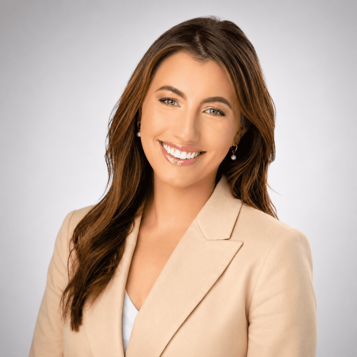 A woman with long brown hair, wearing a beige blazer and white top, smiles at the camera against a plain light gray background.