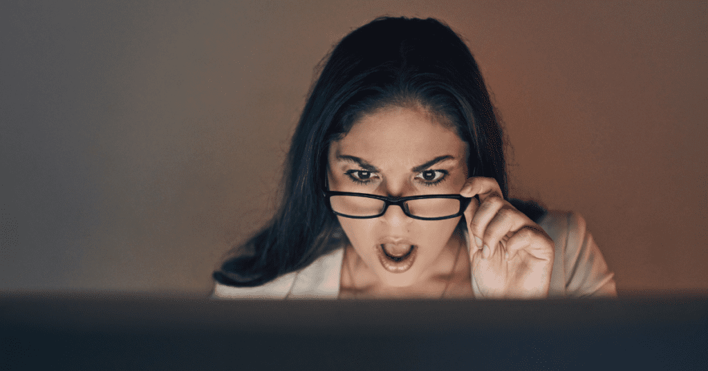 A woman with long dark hair and glasses looks surprised while staring at a computer screen, her mouth open and eyebrows raised. The room is dimly lit, highlighting her face and expression.