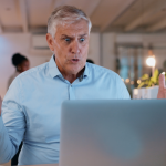 An older man with gray hair wearing a light blue shirt sits at a desk, looking surprised or frustrated at his laptop with his arms raised. The background shows an office setting with people and warm lighting.