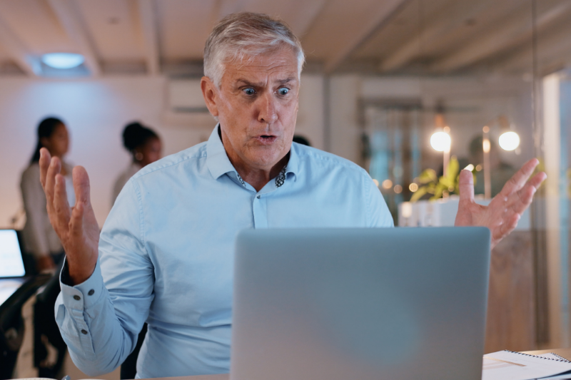 An older man with gray hair wearing a light blue shirt sits at a desk, looking surprised or frustrated at his laptop with his arms raised. The background shows an office setting with people and warm lighting.