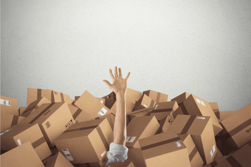 A single hand reaching up from a large pile of cardboard boxes, suggesting the person is buried under the boxes and seeking help.