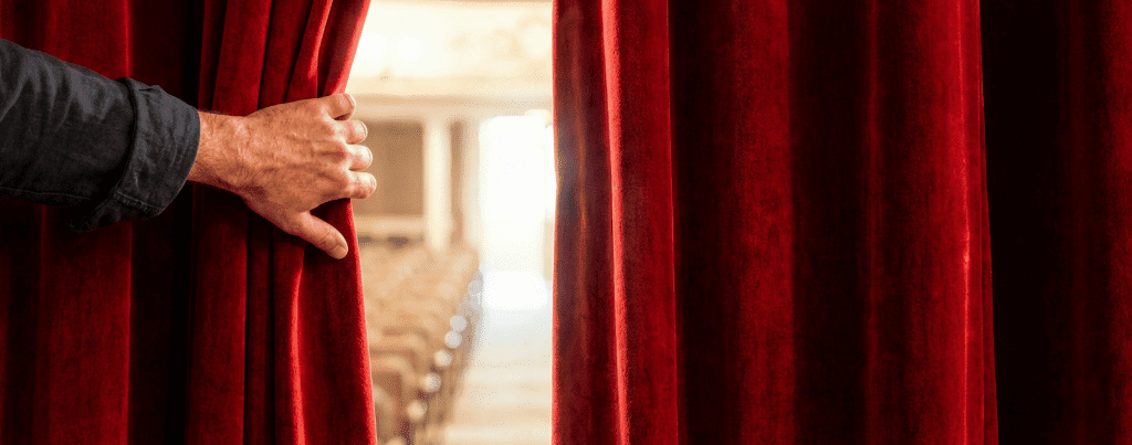 A hand pulls back a red velvet theater curtain, revealing empty rows of seats in an auditorium bathed in soft light.