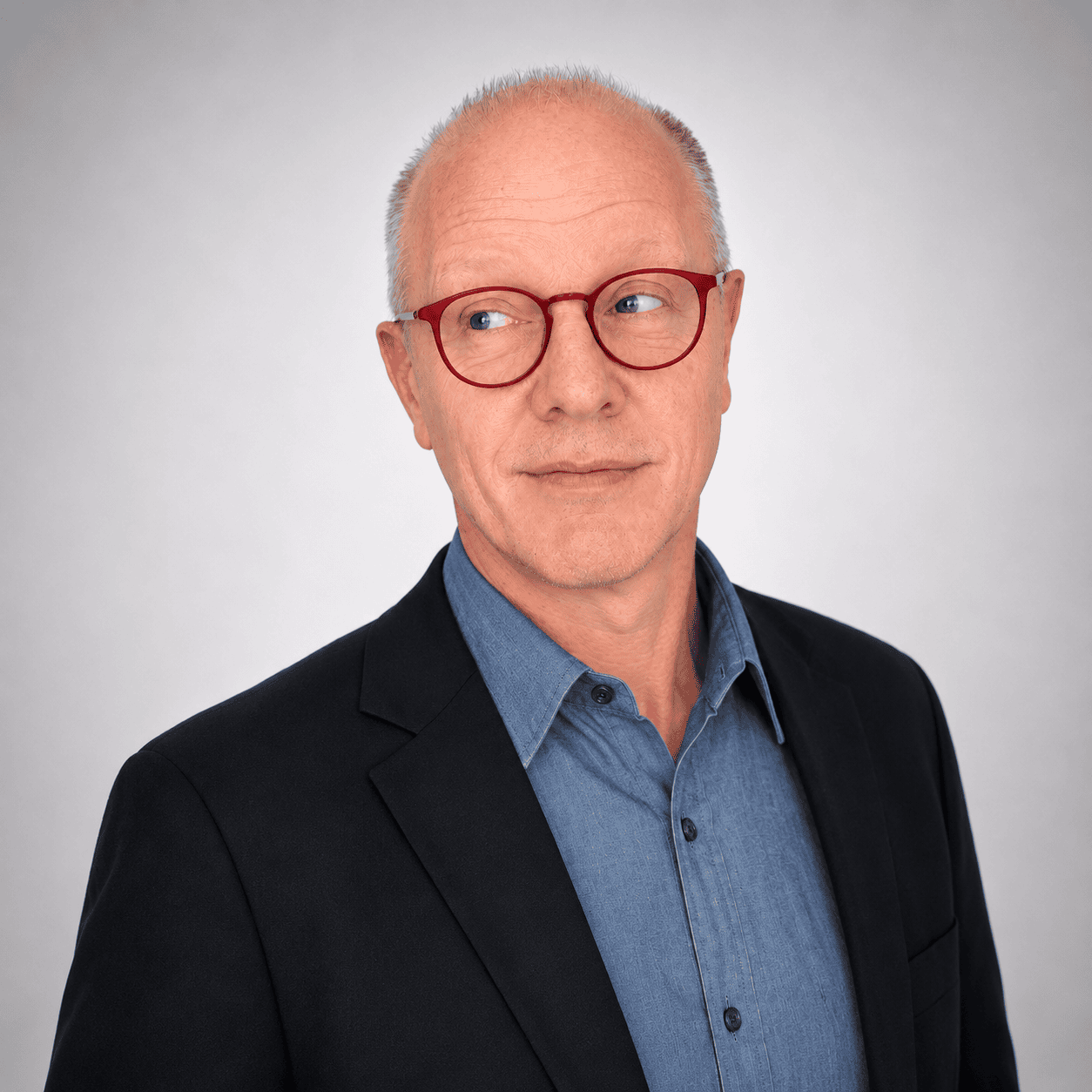 A middle-aged man with short gray hair, red glasses, and a slight smile wears a blue shirt and black blazer. He looks to his left against a plain light gray background.