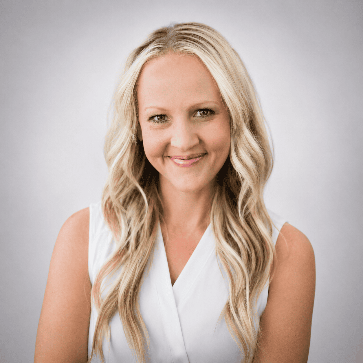 Woman with long, wavy blonde hair and a white sleeveless top smiles at the camera against a light gray background.