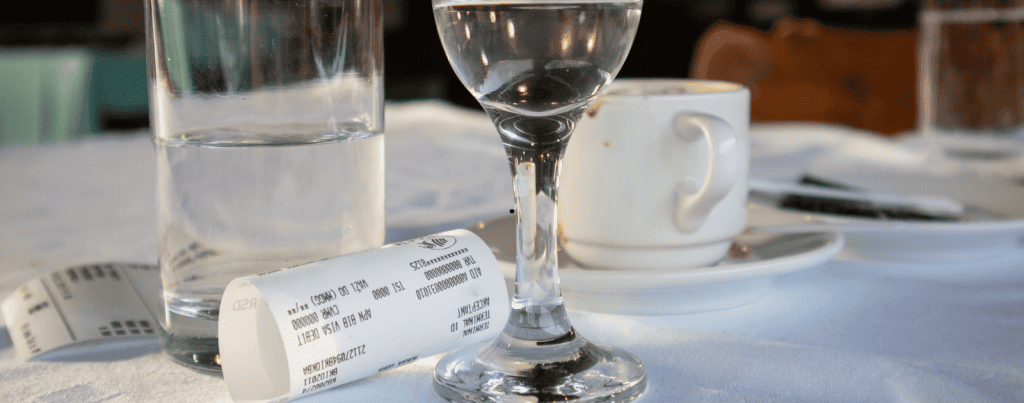 A restaurant table with a glass of water, a coffee cup on a saucer, a small receipt, and another glass. The tablecloth is white and sunlight is shining on the items.